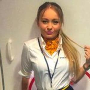 Może przedstawiać: Kobieta w białej koszuli z krótkim rękawem, żółtym szaliku i niebieskiej smyczy. Na koszuli widnieje napis "RYANAIR". Stoi przed białymi drzwiami.
