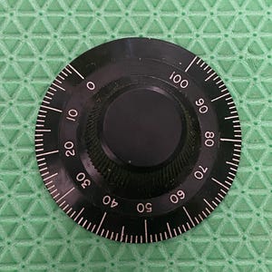 May include: A black rotary dial with white markings and numbers from 0 to 100. The dial has a ridged edge and a smooth, circular centre. The background is a textured green surface.