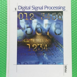 May include: A white book titled "Digital Signal Processing" with a blue and gold design on the cover. The cover features numbers and text, including "1234 5678 9012 3456". The book is on a green surface.