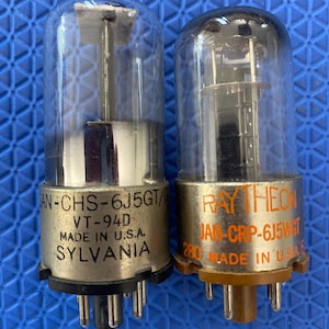 May include: Two vintage vacuum tubes with glass enclosures. One tube is labelled "SYLVANIA" and the other "RAYTHEON". Both tubes have metal bases with pins and are marked "MADE IN U.S.A."