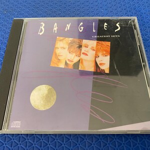 Puede incluir: Una caja de CD con el álbum "Greatest Hits" de The Bangles. La portada muestra el nombre de la banda en una fuente estilizada sobre una foto de los miembros. El fondo es una mezcla de morado y negro con un círculo dorado.