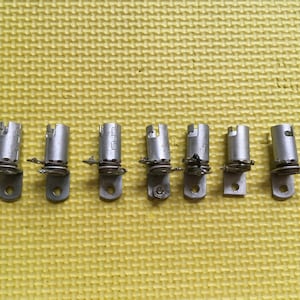 May include: Seven silver-colored metal cylindrical components arranged in a horizontal row. Each piece features a vertical tube with a small side-mounted bracket containing a circular hole, a coiled spring mechanism, and a thin metal wire extension.