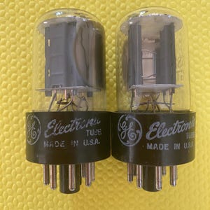 May include: Two clear glass vacuum tubes with internal grey metal components and black bases. Each base features the GE logo, the text "Electronic TUBE" and "MADE IN U.S.A." printed in white. The tubes have multiple metal pins at the bottom.