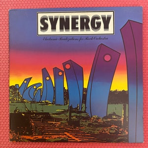 May include: A vintage vinyl record cover with a blue, yellow, and orange color scheme. The cover features the text "SYNERGY" in bold, white letters and the words "Electronic Realizations for Rock Orchestra" in smaller, white letters. The cover art depicts a series of abstract, colorful shapes against a sunset background.