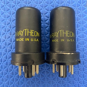 May include: Two black Raytheon vacuum tubes with gold pins. The text "RAYTHEON" and "MADE IN U.S.A." are printed in yellow on the tubes. The tubes have a cylindrical shape with a base that has several pins.
