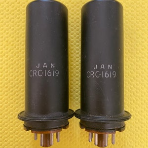 May include: Two black cylindrical vacuum tubes with the text "JAN CRC-1619" printed on them. The tubes have a circular base with several pins. The background is a solid yellow color.