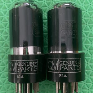 Op de afbeelding: Twee zwarte vacuümbuizen met heldere glazen toppen. De buizen hebben zilveren metalen ringen en zwarte bases met pinnen. De bases zijn gemarkeerd met "GM GENUINE PARTS" en "16V6GT". De achtergrond is groen.