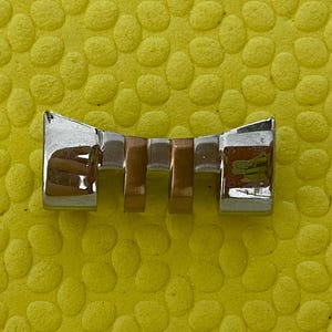 Może przedstawiać: Srebrno-złote ogniwo paska do zegarka o prostokątnym kształcie. Ogniwo ma polerowane wykończenie i mały zielono-żółty emblemat na jednym końcu. Ogniwo jest umieszczone na teksturowanym żółtym tle.