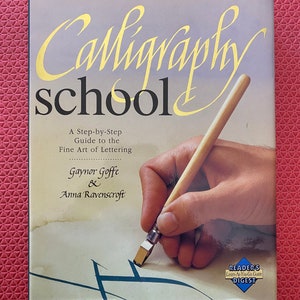 May include: A book cover for "Calligraphy School", a guide to the fine art of lettering. The title is in gold script, with a hand holding a calligraphy pen. Includes the Reader's Digest logo.
