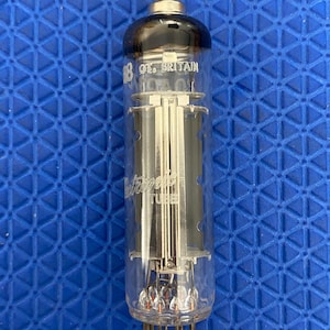 Puede incluir: Una válvula de vacío vintage con un cuerpo de vidrio transparente, componentes internos plateados y una parte superior negra. El tubo tiene una base de metal con pines y el texto "GT. BRITAIN" es visible.