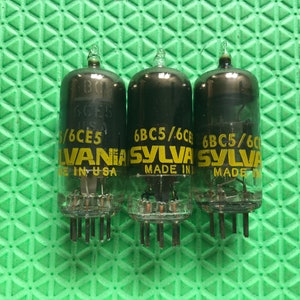 May include: Three vintage vacuum tubes with black glass bodies and clear glass bases. Each tube has the text "6BC5/6CE5" and "SYLVANIA MADE IN USA" printed on it. The tubes are arranged on a green surface.