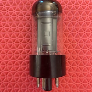 May include: A vintage vacuum tube with a clear glass enclosure and a dark, rounded top. The tube has a black base with several pins extending downward. The internal components are visible through the glass.