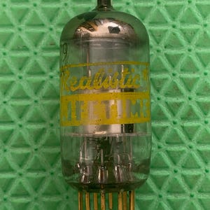 May include: A vintage vacuum tube with a clear glass bulb and gold-colored pins. The tube has a yellow band with the word "Realistic" printed on it. The background is a green textured surface.