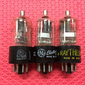 Puede incluir: Tres válvulas de vacío vintage con carcasas de vidrio y bases negras. Las válvulas están etiquetadas con las marcas CBS Hytron, GE Electronics y Raytheon. El texto "MADE IN U.S.A." es visible en cada base. El fondo es una superficie roja texturizada.