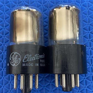 Pode incluir: Dois tubos eletrônicos GE com topos de vidro transparente e bases pretas. O tubo esquerdo apresenta o logotipo GE, o texto Electronic TUBE e MADE IN U.S.A. impressos em branco. Ambos os tubos possuem vários pinos metálicos na parte inferior.