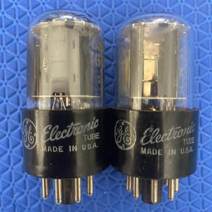 May include: Two vintage GE Electronic Tube vacuum tubes. Each tube has a clear glass bulb and a black base with the GE logo and the text "Electronic Tube MADE IN U.S.A.". The tubes have multiple pins.