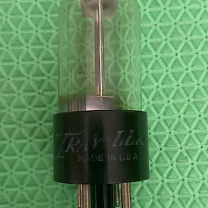 May include: A vintage vacuum tube with a clear glass enclosure and a black base. The base has the text "Trav-Lla" and "Made in U.S.A." The tube has several pins at the bottom and a silver internal structure.