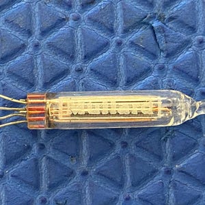 Puede incluir: Un tubo de vacío de vidrio transparente con componentes internos y múltiples cables que se extienden desde un extremo. El tubo tiene forma rectangular con un extremo redondeado. La base es de color marrón rojizo. El tubo está sobre una superficie azul texturizada.