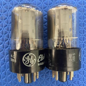May include: Two vintage GE Electronics vacuum tubes. Each tube has a clear glass bulb, a black base with the GE logo, and multiple pins. The text "MADE IN" is visible on the base of one tube, and "4-17 188-5" and "2-48 188-5" are printed on the bases.
