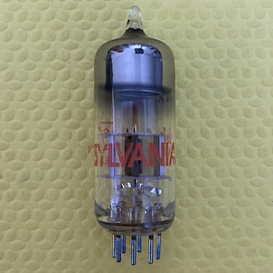 May include: A vintage Sylvania vacuum tube with a clear glass body and a gray top. The word "SYLVANIA" is printed in red on the tube. The tube has multiple pins at the bottom and internal components visible through the glass.