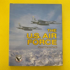 Peut inclure: Couverture de livre "The U.S. Air Force" de Nancy Warren Ferrell. L'image présente deux avions de chasse survolant une chaîne de montagnes. Le titre est en jaune et le logo de la série Lerner's Armed Services est visible.
