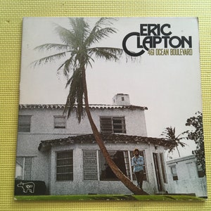 May include: A black and white photo of a house with a palm tree in front of it. The house has a white exterior and a tiled roof. The palm tree is tall and has a thick trunk. The photo is titled "Eric Clapton 461 Ocean Boulevard".