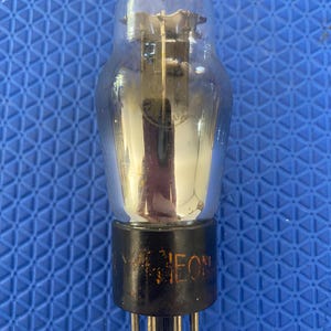 May include: A vintage vacuum tube with a clear glass bulb and a black base. The base has the word "NEON" printed on it. The tube has several pins at the bottom and internal components visible through the glass.
