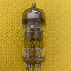 May include: A clear glass vacuum tube with a silver internal structure and a series of pins at the base. The tube has the text "6DJ7/EF84" and "SYLVANIA" printed in yellow. The top of the tube is rounded.