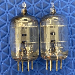 May include: Two vintage vacuum tubes with clear glass bulbs and gold-colored pins. The internal components are visible, including filaments and support structures. The tubes have a cylindrical shape with a pointed top.