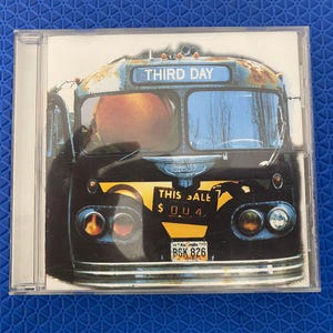 Könnte beinhalten: Eine CD-Hülle mit der Grafik eines Oldtimer-Busses mit blauem Zielschild mit der Aufschrift THIRD DAY. Der Bus ist schwarz und gelb mit einem Schild mit der Aufschrift THIS SALE $ 0.0.4 und einem Georgia-Nummernschild B6K 826. Eine orangefarbene Kugel ist zu sehen.