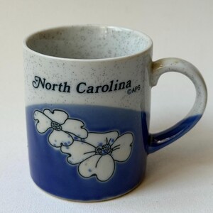 May include: A ceramic mug with a blue and white speckled design. The mug features the words "North Carolina" in black, along with a blue band and white floral illustrations. The handle is also blue.