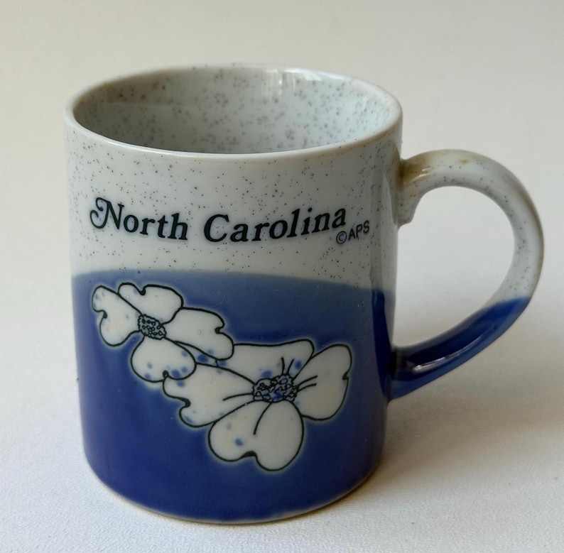 May include: A ceramic mug with a blue and white speckled design. The mug features the words "North Carolina" in black, along with a blue band and white floral illustrations. The handle is also blue.