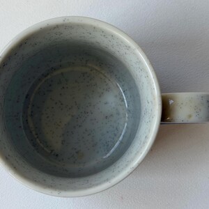 May include: Overhead shot of a speckled ceramic mug. The mug is light grey with darker grey speckles. The handle is a lighter shade of grey. The interior of the mug is a darker grey.