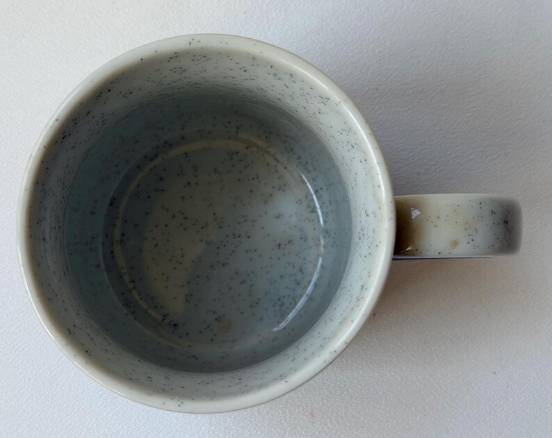 May include: Overhead shot of a speckled ceramic mug. The mug is light grey with darker grey speckles. The handle is a lighter shade of grey. The interior of the mug is a darker grey.