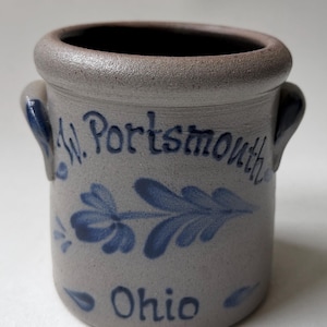 May include: A cylindrical stoneware crock with a thick rim and two side handles. The gray body features blue hand-painted floral motifs and the blue text W. Portsmouth Ohio. The vessel has a textured, matte finish and a wide, open top opening.