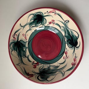 May include: A decorative ceramic bowl with a cream background and a red rim. The bowl features a central red circle surrounded by a green ring. The cream surface is adorned with black and green floral designs and pink accents.
