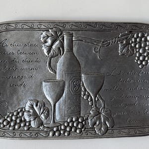Puede incluir: Una bandeja rectangular de color peltre con imágenes en relieve con temática de vino. La bandeja presenta una botella de vino, dos copas de vino, uvas y hojas. El texto está en relieve en la bandeja, incluyendo frases sobre amistad y vino.