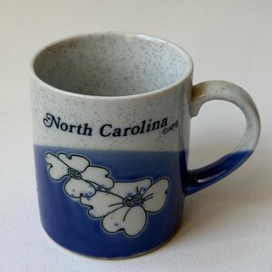 May include: A ceramic mug with a speckled white and blue upper section and a solid blue lower section. The mug has the words "North Carolina" in black script and white floral designs. The handle is blue.