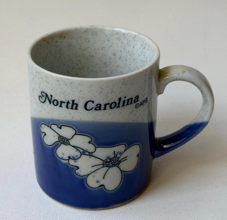 May include: A ceramic mug with a speckled white and blue upper section and a solid blue lower section. The mug has the words "North Carolina" in black script and white floral designs. The handle is blue.