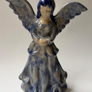 May include: A ceramic angel figurine featuring a glossy finish with a mottled blue and cream glaze. The figure has outstretched wings, dark blue hair, and a long, flowing gown with a flared base, standing in a neutral, upright posture.