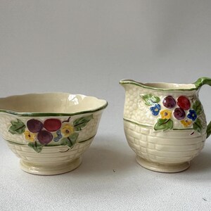 May include: A vintage cream-colored sugar bowl and creamer set with a woven basket design and floral accents. The set features a green rim and colorful flowers and berries.