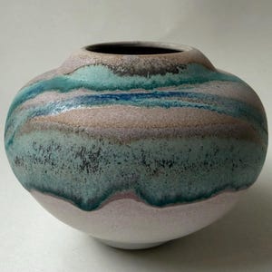 May include: A round ceramic vase with a wide, bulbous body and a narrow opening. The surface features a layered glaze with horizontal bands of muted pink, soft mint green, and deep teal, with a speckled, textured finish across the midsection.