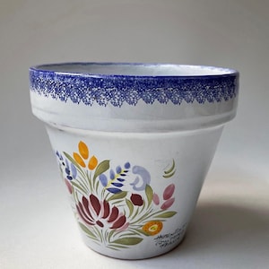 May include: White ceramic flower pot with a blue rim and a floral design in shades of red, pink, yellow, blue, and green. The pot is hand-painted and has the text "Atelier du Quai" and "France" on the bottom.