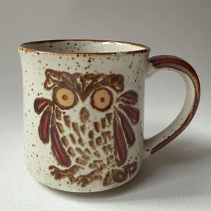 May include: A ceramic mug with a speckled off-white glaze featuring a hand-painted brown owl design. The owl has large yellow eyes and dark brown wing details. The mug has a brown rim and a matching brown handle, with a cylindrical shape.