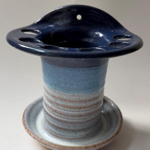 May include: A ceramic toothbrush holder with a dark blue glazed top featuring multiple circular openings and a small hole for wall mounting. The cylindrical body transitions from a light blue glaze to a white and brown striped base.