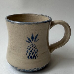 Könnte beinhalten: Beige Keramikbecher mit blauem Ananas-Design. Der Becher hat eine geschwungene Form, einen Henkel und einen blauen Rand. Die Ananas ist eine stilisierte Illustration. Geeignet für heiße oder kalte Getränke.