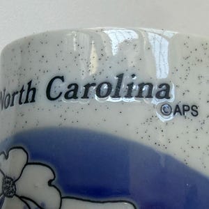 May include: Close-up of a ceramic mug with the words "North Carolina" in black lettering. The mug has a speckled white background with a blue floral design and a blue band. The APS logo is also visible.