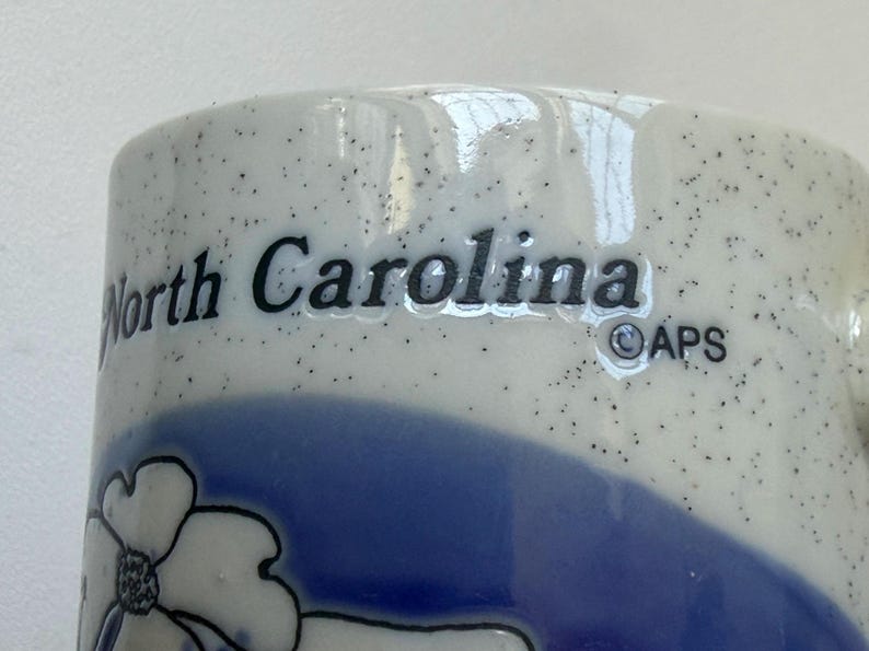May include: Close-up of a ceramic mug with the words "North Carolina" in black lettering. The mug has a speckled white background with a blue floral design and a blue band. The APS logo is also visible.
