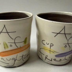 May include: Two off-white ceramic cups with dark brown interiors. The left cup features the text A cup of laughter with an orange brushstroke. The right cup features the text A cup of friends with yellow and purple brushstrokes in a hand-painted style.