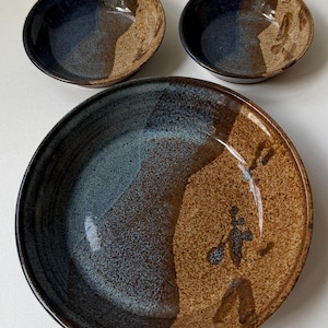 May include: A set of three ceramic bowls. The bowls have a two-tone design, with a dark blue-grey glaze on one side and a tan speckled glaze on the other. The set includes two smaller bowls and one larger bowl.